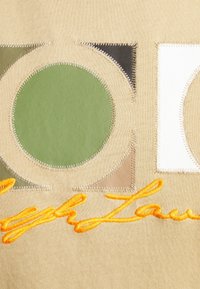 Béžová tkanina má vícebarevné kulaté nášivky v zelené, černé a bílé barvě, přičemž na ní je vyšitý text "Yves Laurent" v oranžové barvě.