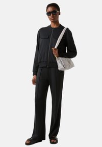 Zwarte bomberjack met ritssluiting en twee borstzakken, gecombineerd met bijpassende zwarte wijde broekspijpen en robuuste zwarte sandalen. Grijze gewatteerde handtas.