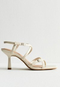 Beige remsknit högklackad sandal med en glansig yta, justerbara spännen och en lätt texturerad innersula. Har en smal, avsmalnande klack.