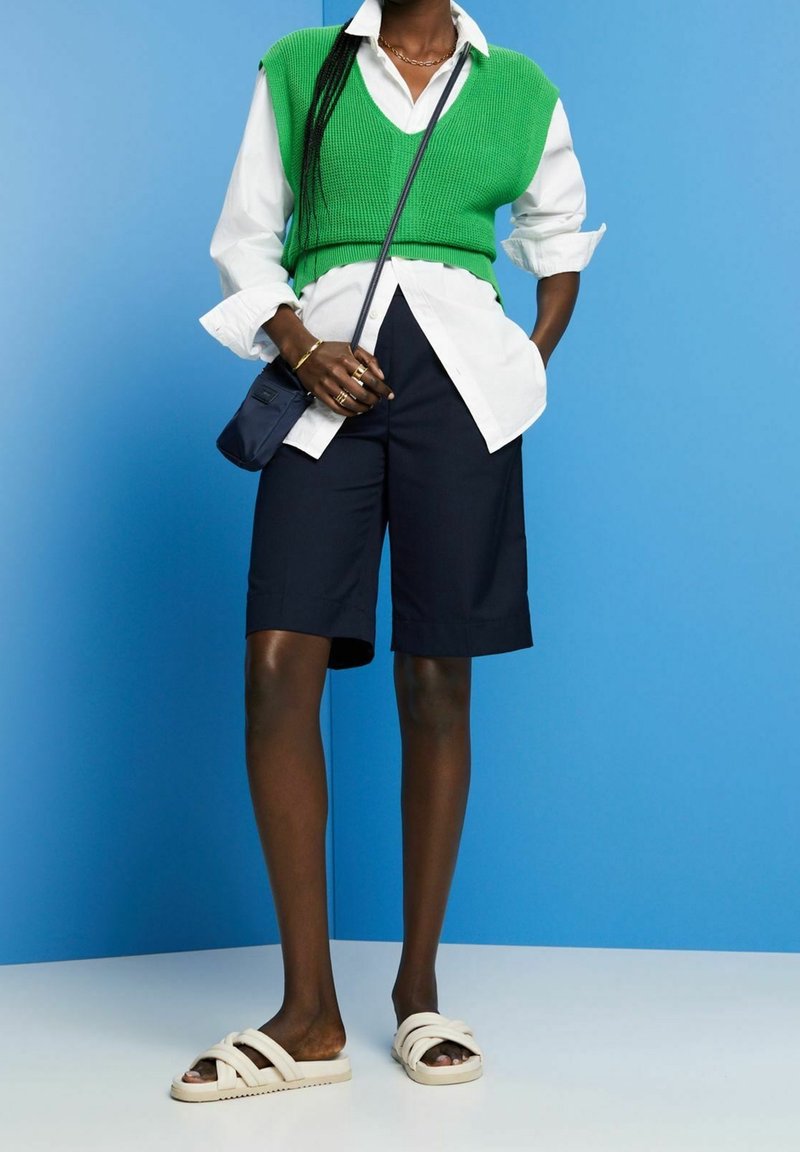 Personne portant un short long noir, une chemise blanche boutonnée, un gilet tricoté vert, des sandales beige, et portant un petit sac bandoulière noir.