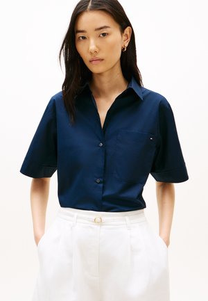 Mladá žena s dlhými tmavými vlasmi, oblečená v tmavomodrej krátkorysovej košeli na gombíky a bielych vysokých nohaviciach, ruky v vreckách.