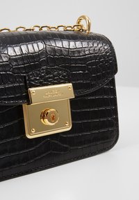 Svart läderhandväska med krokodilprägling, gold hardware-lås och kedjespänne. Har en rektangulär form och en texturerad yta.
