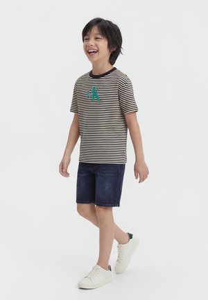 Γελαστό αγόρι που περπατά φορώντας μαύρο και λευκό ριγέ μπλουζάκι με πράσινο λογότυπο, σκούρα τζιν σορτς και λευκά αθλητικά παπούτσια.