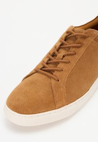 Svijetlosmeđe suede tenisice s glatkom teksturom, okruglim vrhom, šivenim detaljima i smešnim vezicama, s bijelim gumiranim potplatom.
