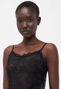 Camisole en dentelle noire avec de fines bretelles, présentant un décolleté festonné et des motifs floraux complexes. Texture douce, conception légère.