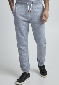 Grå melange joggebukser laget av mykt stoff, med snøring i elastisk liv og sidelommer, sammen med svarte joggesko.