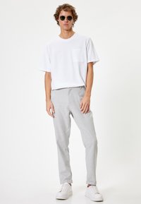 Hvid bomulds T-shirt med en forlommes, parret med lysegrå bukser. Model iført runde sorte solbriller og hvide sneakers.