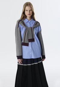 Blauw gestreepte blouse met knopen, gecombineerd met een grijze trui om de schouders geknoopt, gedragen met een zwarte plissérok met kantafwerking.
