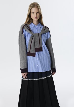 Blauw gestreepte blouse met knopen, gecombineerd met een grijze trui om de schouders geknoopt, gedragen met een zwarte plissérok met kantafwerking.