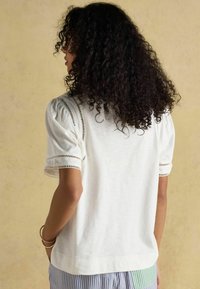 Valge lühikeste varrukatega pluus, millel on puffitud õlad ja lõikeaukude detailid varrukate ääres. Pehme kangas ja mugav lõige.