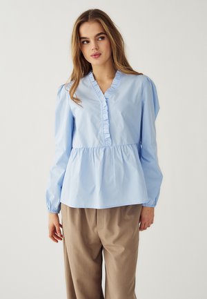 Frau trägt eine hellblaue, mit Rüschen versehene Bluse mit V-Ausschnitt und helle, beige Hosen mit weitem Bein, steht vor einem schlichten, neutralen Hintergrund.