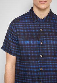 Korte mouwen overhemd met knopen in donkerblauwe stof met een geruit patroon van zwarte en blauwe lijnen, met bruine knopen en een puntige kraag.