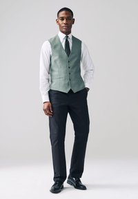 Gilet vert texturé porté sur une chemise blanche, associé à un pantalon noir et des chaussures noires brillantes. Le gilet est orné de boutons et la cravate présente de petits points.