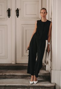 Woman in black sleeveless top and pants leans against white wall on stone steps, holding beige handbag and wearing white pointed shoes.