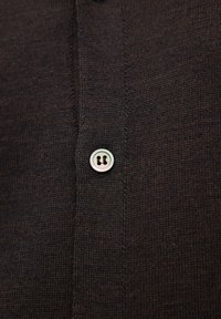 Brązowa dziana tkanina z zapięciem na guzik, z okrągłym, opalizującym guzikiem. Widoczny teksturowany wzór dzianiny oraz detale przeszycia.