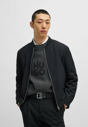 Black bomber jacket with knit collar, paired with a textured black sweater featuring a bold logo, and tailored black trousers with a belt.