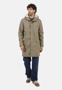 Manteau imperméable kaki avec une capuche douce en fausse fourrure, des boutons-pression et une coupe décontractée. Associé à un jean bleu et des baskets marron.