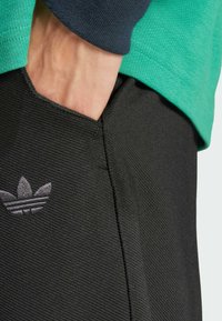 Crne kratke hlače s teksturiranom tkaninom i malim sivoim Adidas logom. Hlače imaju v-oblik rez na šavu džepa. Vidljiva zelena i tamnoplava majica.