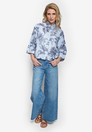 Blonde vrouw met een blauw gestreept bloemenhemd, wijde blauwe spijkerbroek en bruine sandalen, staand met haar handen bij haar heupen.