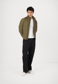 Veste vert olive avec un col haut et une fermeture éclair à l'avant, associée à un t-shirt blanc et un pantalon cargo noir ; le mannequin porte des baskets blanches.