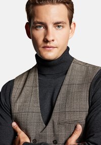 Brązowa kamizelka w kratę noszona na czarnym swetrze z golfem. Kamizelka ma niski dekolt oraz podwójne zapięcie na guziki, a jej powierzchnia ma fakturowane wykończenie.