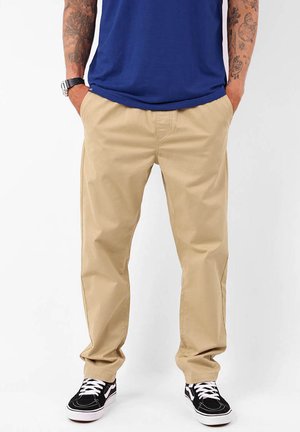 Chinos kaki avec une coupe droite, fabriqués en tissu mélangé de coton ; dotés de poches latérales et portés avec des baskets noires et une chemise bleue.