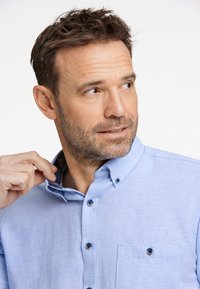 Ljust blå skjorta med knappar gjord av tyg med texturerad yta. Har en bröstficka och kontrasterande detaljer på knapparna.