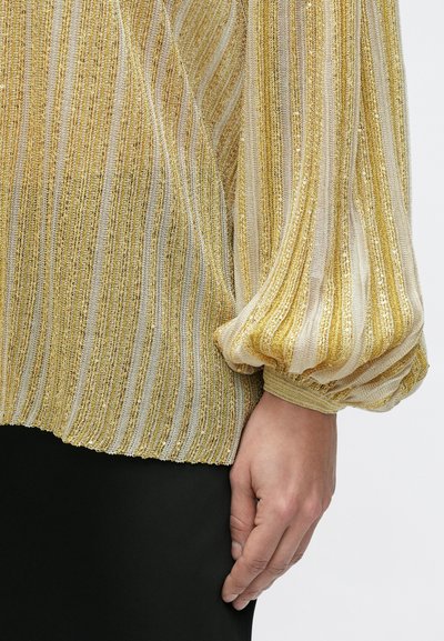 Hånd hvilende ved siden iført langærmet top med gyldne og hvide lodrette striber samt subtile palietter, kombineret med sort bund.