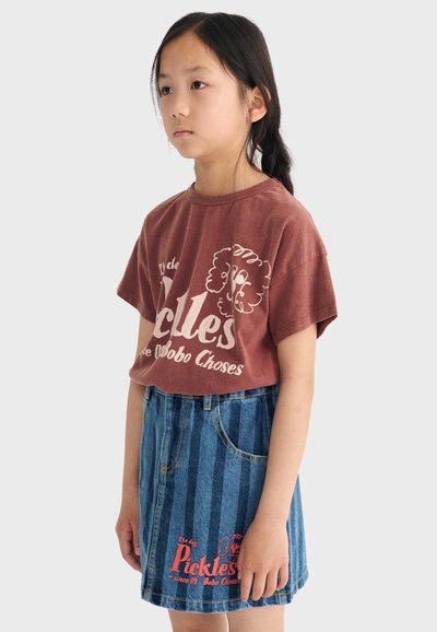 Ragazza giovane con capelli lunghi e scuri, indossa una maglietta marrone con stampa grafica e una gonna di denim a righe blu con scritte rosse, in piedi di profilo.