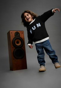 Dijete u crnom džemperu s natpisom "FUN" svijetloplave boje, plavim trapericama i bež tenisicama, stoji pored drvenog zvučnika s dva crna zvučnika.