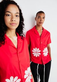 Červené tričká s krátkymi rukávmi a bielou kvetinovou výšivkou v oblasti pása, skombinované s čiernymi nohavicami s vlnitými bielymi akcentmi.