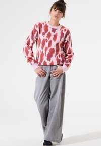 Suéter rosa con manchas de leopardo rojas y burdeos, textura de punto, puños acanalados. Combinado con unos pantalones grises holgados y zapatos negros.