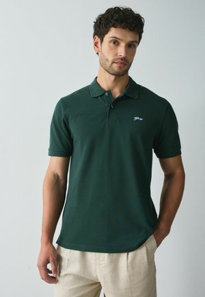 Mand iført en mørkegrøn kortærmet polo med en lille logo på brystet og beige bukser, der står mod en ensfarvet baggrund.
