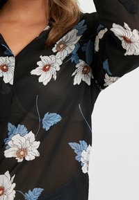 Sort gennemsigtig bluse med hvidt og blåt blomstermønster. Har krave og knapper, og er lavet af et let, struktureret stof.