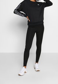 Czarna bluza z logo na klatce piersiowej w białym kolorze i wzorzystymi rękawami, zestawiona z czarnymi leginsami z białymi paskami po bokach. Uzupełniona o białe sneakersy.