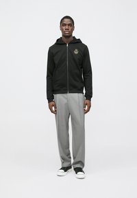 Svart hettejakke med glidelås og logo foran, grå skreddersydde bukser og svarte joggesko. Modellen står rett opp og ned mot en ensfarget bakgrunn.