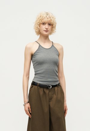 Νεαρό άτομο με σγουρά ξανθά μαλλιά που φοράει γκρι αμάνικο τοπ, καφέ φαρδιά παντελόνια, μαύρη ζώνη και ασημένιο βραχιόλι, στέκεται μπροστά σε απλό φόντο.