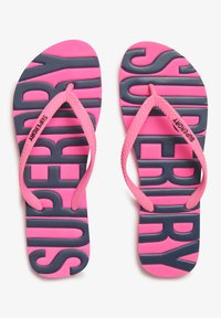 Roze rubberen teenslippers met marineblauwe "SUPERDRY" letters op de textuur van de voetbed en bijpassende roze bandjes. Plat zoolontwerp.
