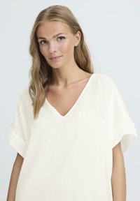 Top bianco sporco con scollatura a V e maniche corte svasate, caratterizzato da una texture a coste e una vestibilità rilassata. Design semplice e minimalista.