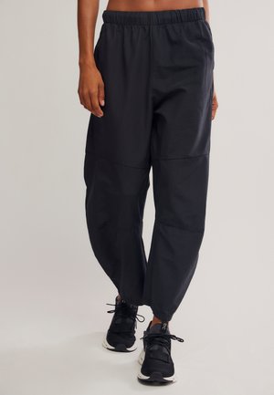 Personne portant un pantalon noir ample avec une taille élastique et des chaussures de sport noires, debout devant un fond uni.