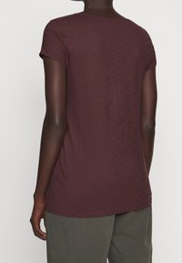 Viininpunainen t-paita, jossa on pyöreä kaula-aukko ja lyhyet hihat, pehmeästä kankaasta valmistettu, vartalonmyötäinen malli ja hieman pidempi takasivu.