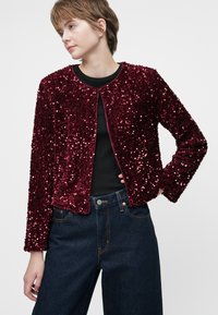 Sarkana zvīņota jaka ar apaļu kakla izgriezumu un garām piedurknēm, ar strukturētu apdari un īsu dizainu, apvienota ar tumšiem džinsu biksēm.