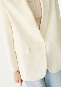 Kremowa marynarka o gładkiej fakturze, z pojedynczym zapięciem na guzik i dwoma przednimi kieszeniami, noszona na beżowej bluzce o subtelnym wzorze.