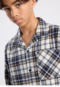 Plaid shirt in shades of navy, cream, and brown in flannel material. Features a button front, collar, and chest pocket with diagonal pattern.