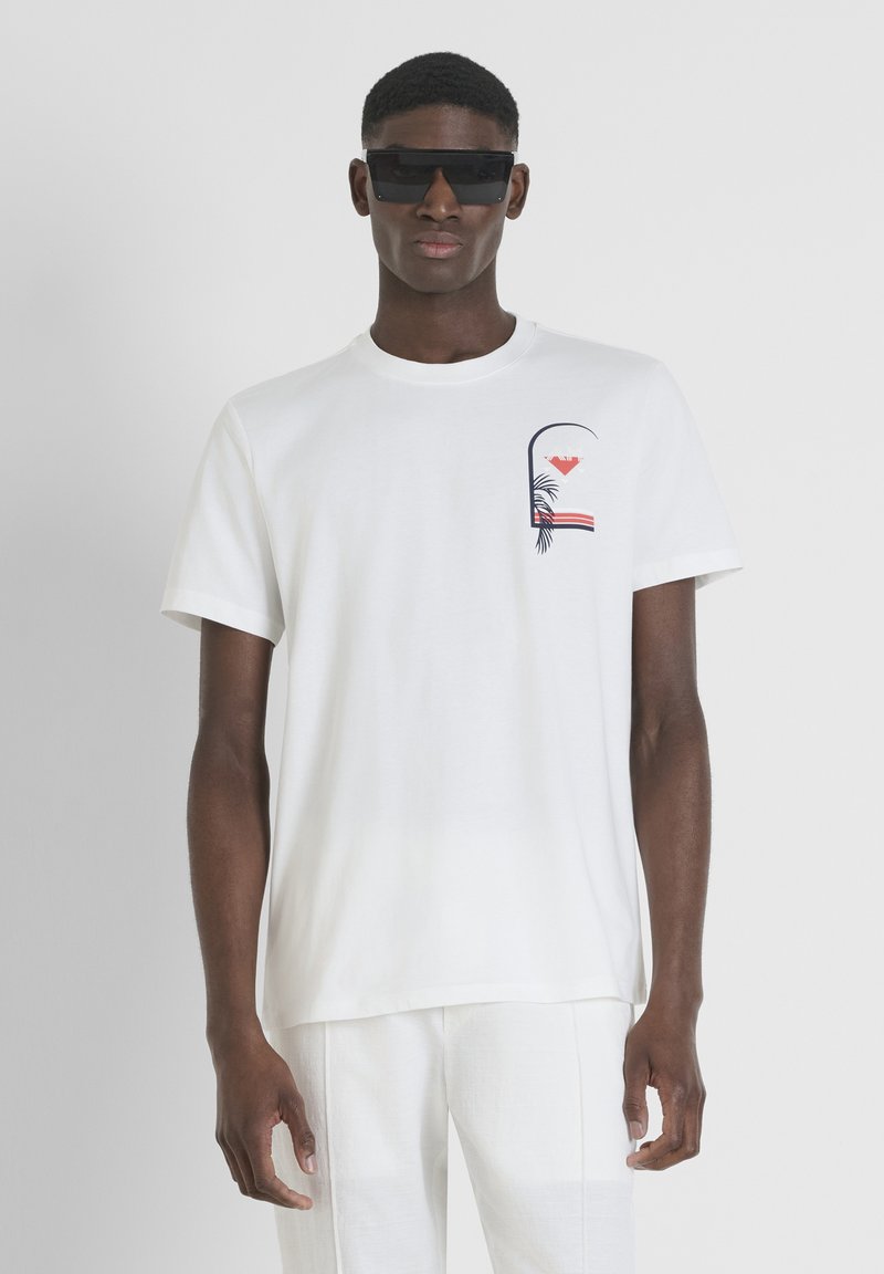 Camiseta blanca de algodón con un pequeño diseño impreso de hojas de palma y rayas en el lado izquierdo del pecho. Modelo con gafas de sol oscuras.