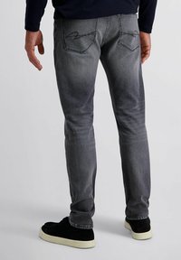 Homme portant un jean gris pâle et des baskets noires avec des semelles blanches, se tenant contre un fond clair.