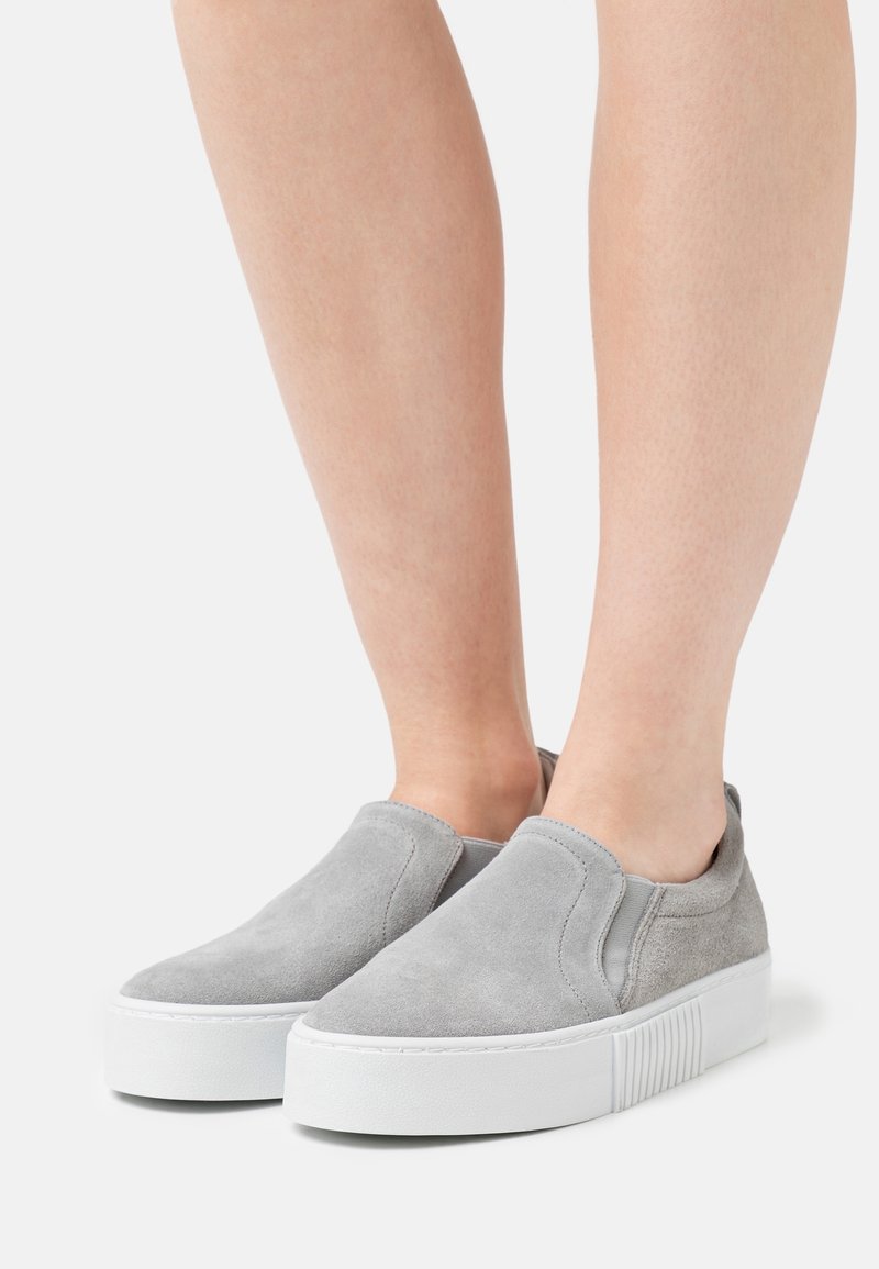 Pieds portant des baskets à enfiler en daim gris clair avec des semelles épaisses blanches sur fond blanc.