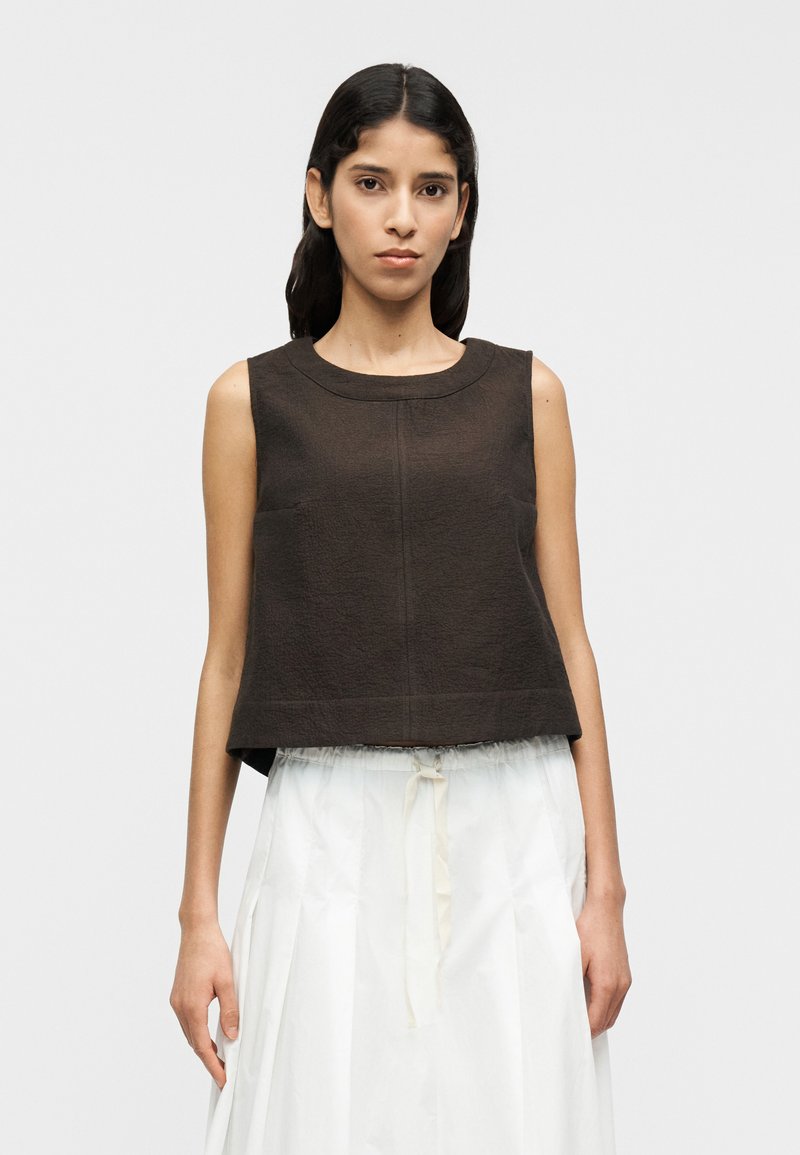 Καφέ αμάνικη μπλούζα με υφή και στρογγυλή λαιμόκ οψη, φορεμένη πάνω από λευκή αέρινη φούστα με κορδόνι στη μέση.