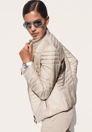 Beige gewatteerde jas met een hoge kraag, ritsen en een gladde textuur. Draag met reflecterende zonnebril en een witte horloge.