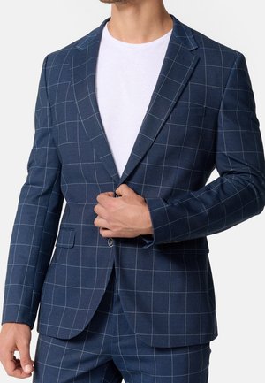 Mann befestigt Knopf an marineblauem Anzug mit weißem Fensterkaromuster, trägt darunter ein schlichtes weißes T-Shirt.